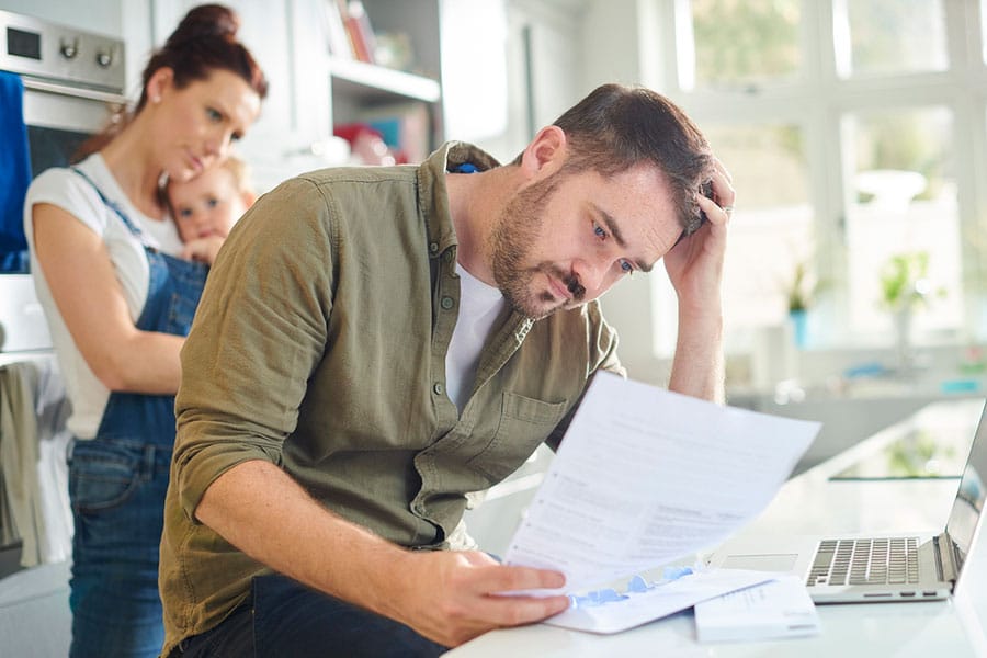 A man sits at a kitchen table, anxiety etched on his face as he reads papers. A woman holding a child stands in the background, both looking concerned. A laptop and bills sit on the table in a bright kitchen.