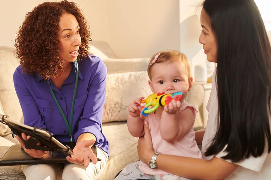 A woman with curly hair speaks to another woman holding a baby. The baby holds a colorful toy and looks toward the camera. Seated on a sofa in a well-lit room, they discuss mindfulness therapy as the woman speaking holds a tablet.