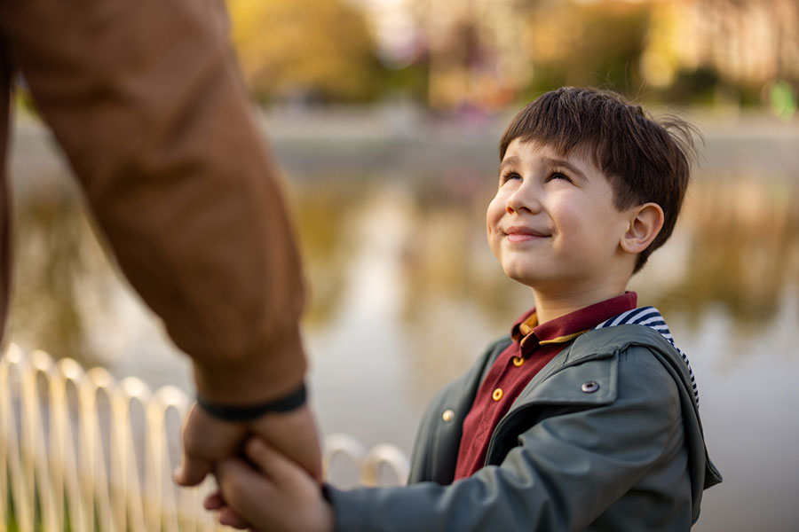 A young boy smiles and looks up while holding hands with an adult outdoors near a body of water, capturing a joyful moment in their adoption journey, with blurred trees and a fence in the background.