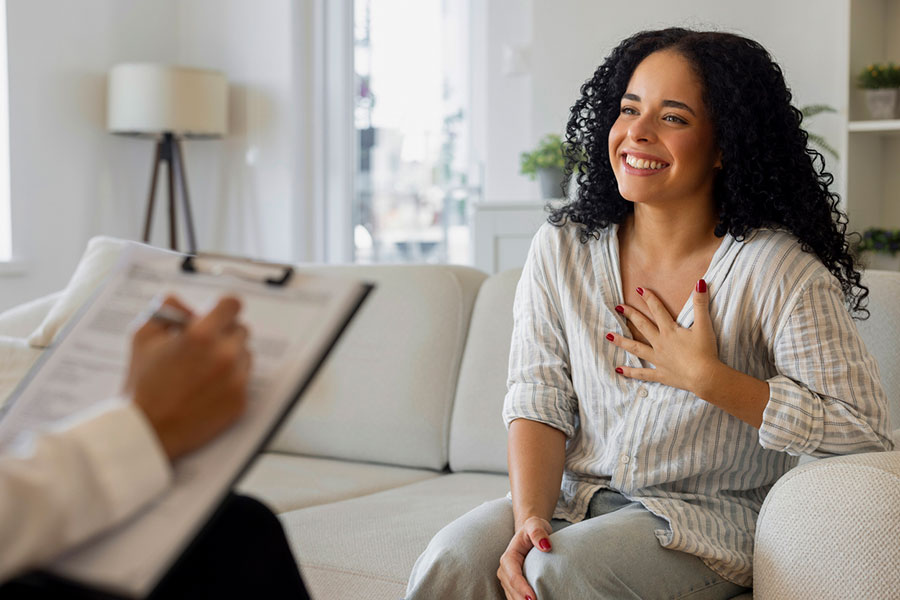 A smiling woman sits on a couch with her hand on her chest, talking to a therapist who is holding a clipboard and taking notes in a bright, comfortable room—breaking the stigma around mental health through open conversation.