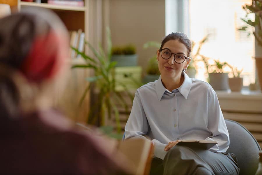 A therapist with glasses and a clipboard sits in an office, attentively listening to another person whose back is facing the camera. There are plants and a window in the background, creating a warm atmosphere ideal for mental health discussions.
