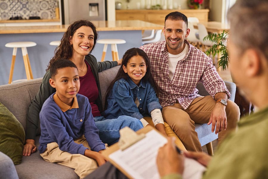 A smiling family of four sits together on a couch, facing a person with a clipboard, possibly in a counseling or therapy session, in a bright modern living room—highlighting the benefits of therapy for mental health and family well-being.