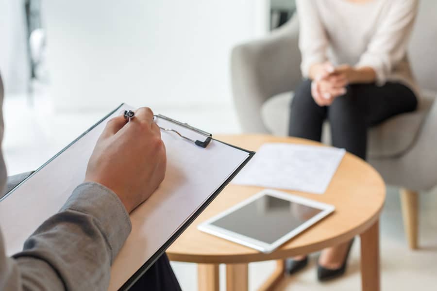 A person holding a clipboard and pen takes notes while another person sits across from them in a chair beside a table with papers and a tablet, illustrating the supportive environment often found in therapy sessions focused on mental health.