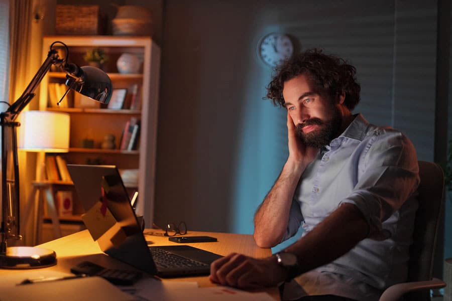 Man with curly hair and beard sits at a desk at night, smiling while looking at his laptop. The room is dimly lit, with a desk lamp, shelves, and a clock on the wall in the background, creating a calm atmosphere that suggests he might be exploring resources about mental health or discovering the benefits of therapy online.