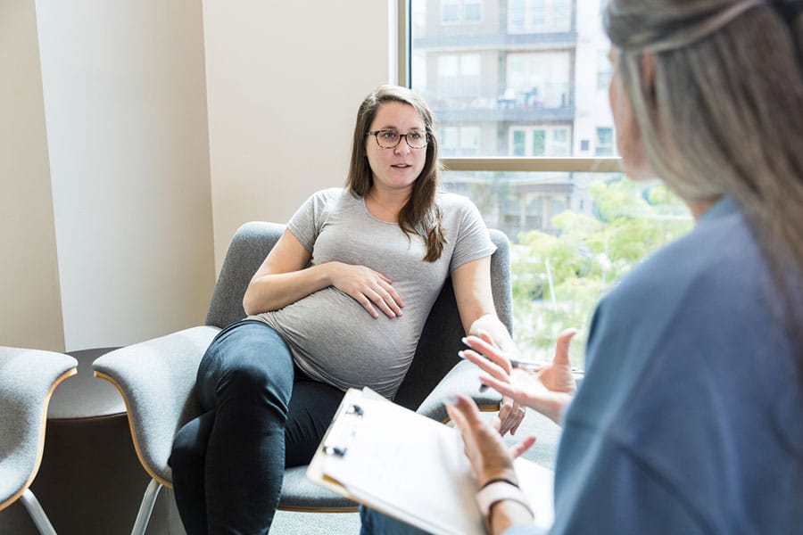 A pregnant woman sits in a chair, holding her belly, while talking to a healthcare professional who holds a clipboard in a brightly lit office, highlighting the importance of maternal support during pregnancy.