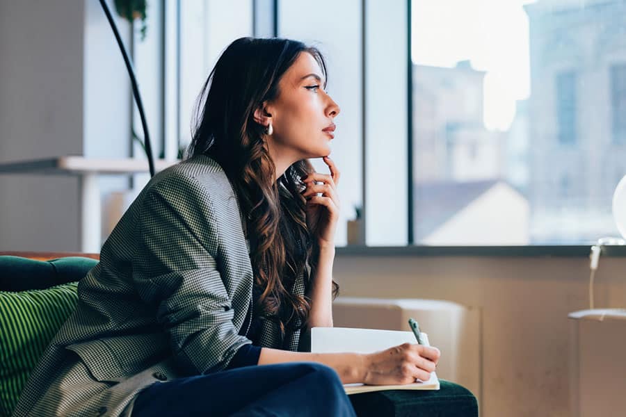 A woman sits indoors by a large window, holding a notebook and pen, looking thoughtful and contemplative, as if she is looking within herself to find inspiration and clarity.