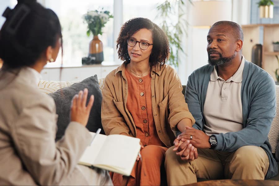 A couple sits close together, holding hands, while talking with a professional who holds a notebook. The setting appears to be a comfortable, well-lit office or living room, providing a supportive environment for therapy focused on navigating different life stages.
