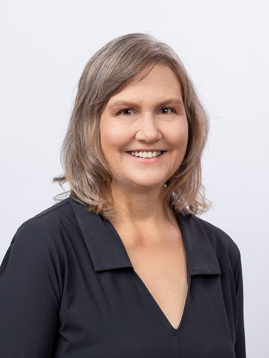A middle-aged woman with shoulder-length light brown hair smiles at the camera. She is wearing a black blouse with a V-neckline. The background is plain white.