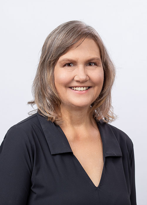 A woman with shoulder-length gray hair and a friendly smile wears a black blouse with a wide collar, standing against a plain white background.